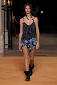 Model walking a runway in a black beaded slip dress with a blue floral fringe hem and black ankle boots.