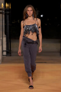 Model walking a runway at night in a silver beaded fringe crop top and gray draped pants with a studded belt.