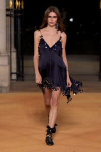 Model on runway wearing a navy mini dress with sequin pom-poms and a fringed, scalloped hem, paired with black lace-up shoes.