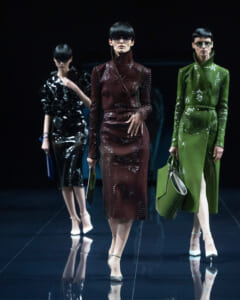 Three models walk a glossy runway wearing high-neck, knee-length, metallic dresses in burgundy, black, and green with oversized sunglasses and matching accessories.