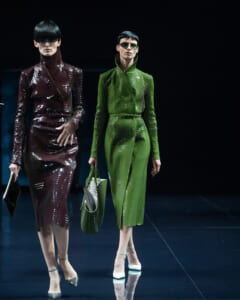 Two models walk a runway in glossy, high-fashion outfits: one in burgundy sequined suit with sunglasses, the other in a bright green matching coat-and-skirt set, carrying a green bag and wearing white heels.