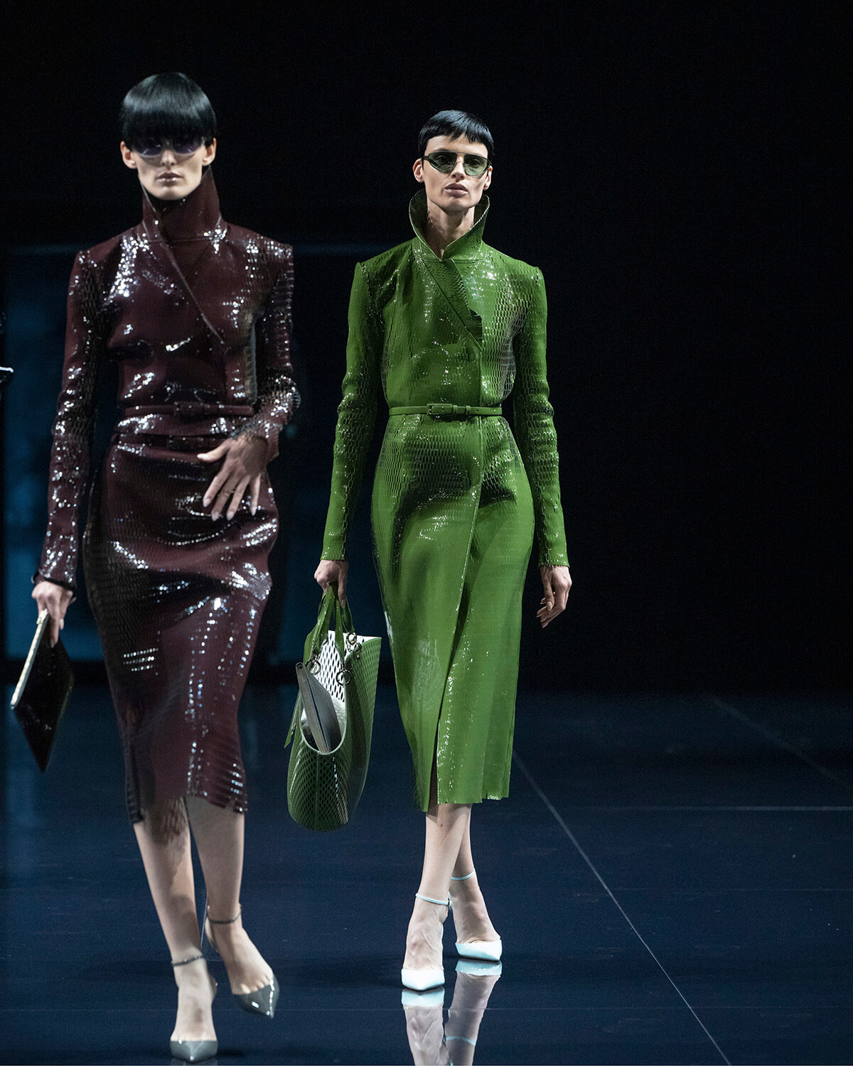 Two models walk a runway in glossy, high-fashion outfits: one in burgundy sequined suit with sunglasses, the other in a bright green matching coat-and-skirt set, carrying a green bag and wearing white heels.