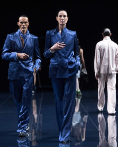 Two models walk a fashion runway in matching cobalt blue suits under dark lighting, glossy floor reflecting their outfits
