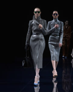 Two models walk a fashion runway in metallic, patterned suits with high collars and sunglasses; blue and silver tones reflect on the glossy floor.
