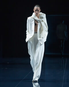 Runway model walking in an oversized white suit with a draped scarf, black bra visible beneath the blazer