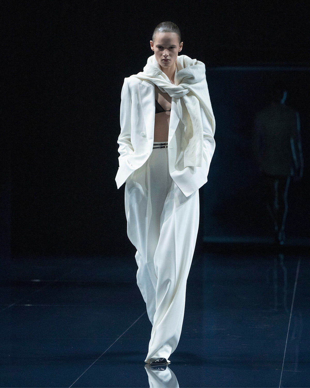 Runway model walking in an oversized white suit with a draped scarf, black bra visible beneath the blazer