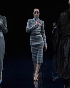 Model walking a runway in a metallic silver patterned dress with a high collar and blue sunglasses. (Front view)