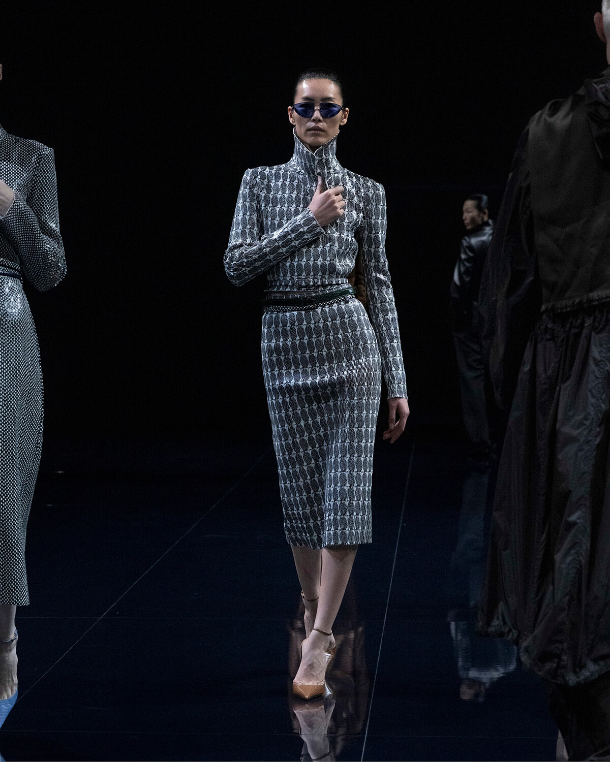 Model walking a runway in a metallic silver patterned dress with a high collar and blue sunglasses. (Front view)
