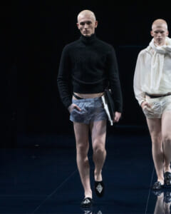 Male model walks the runway in a black knit turtleneck and gray shorts, holding a folder; another model in white outfit stands nearby.
