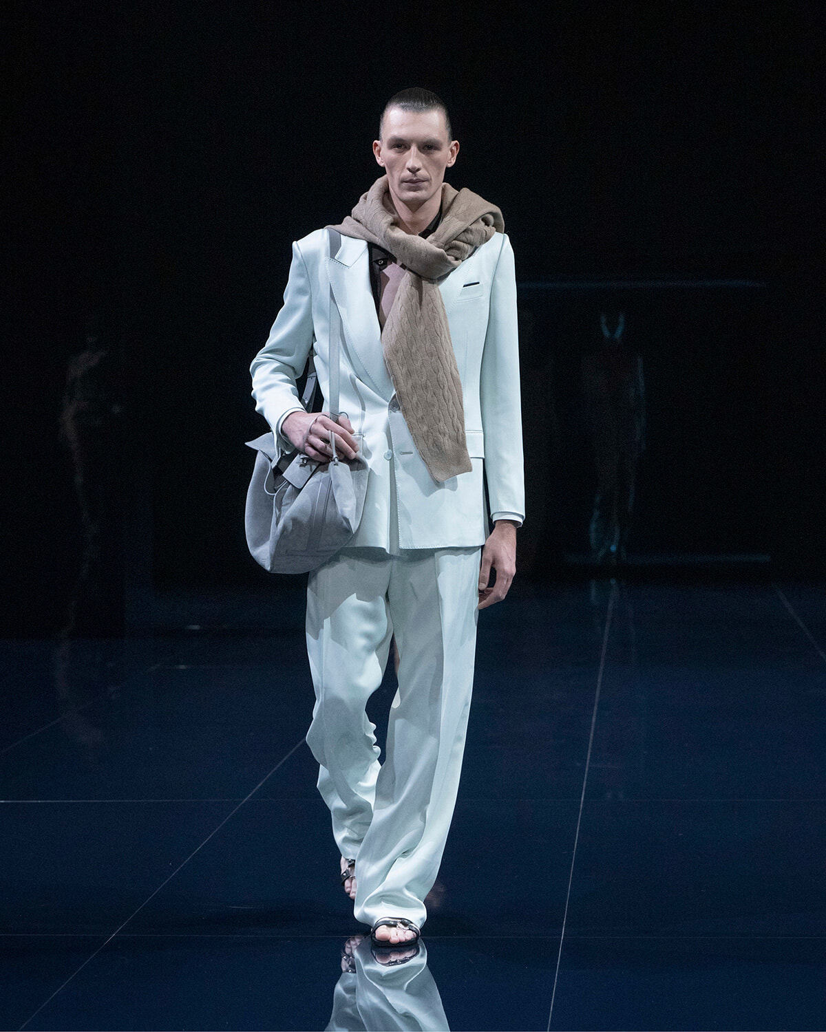 Male model walking a fashion runway in a pale blue suit with a beige scarf and gray bag.