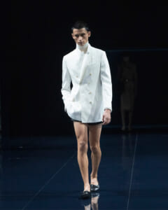 Male model walks a fashion runway wearing a white double-breasted blazer, white turtleneck, and black shorts on a dark stage.