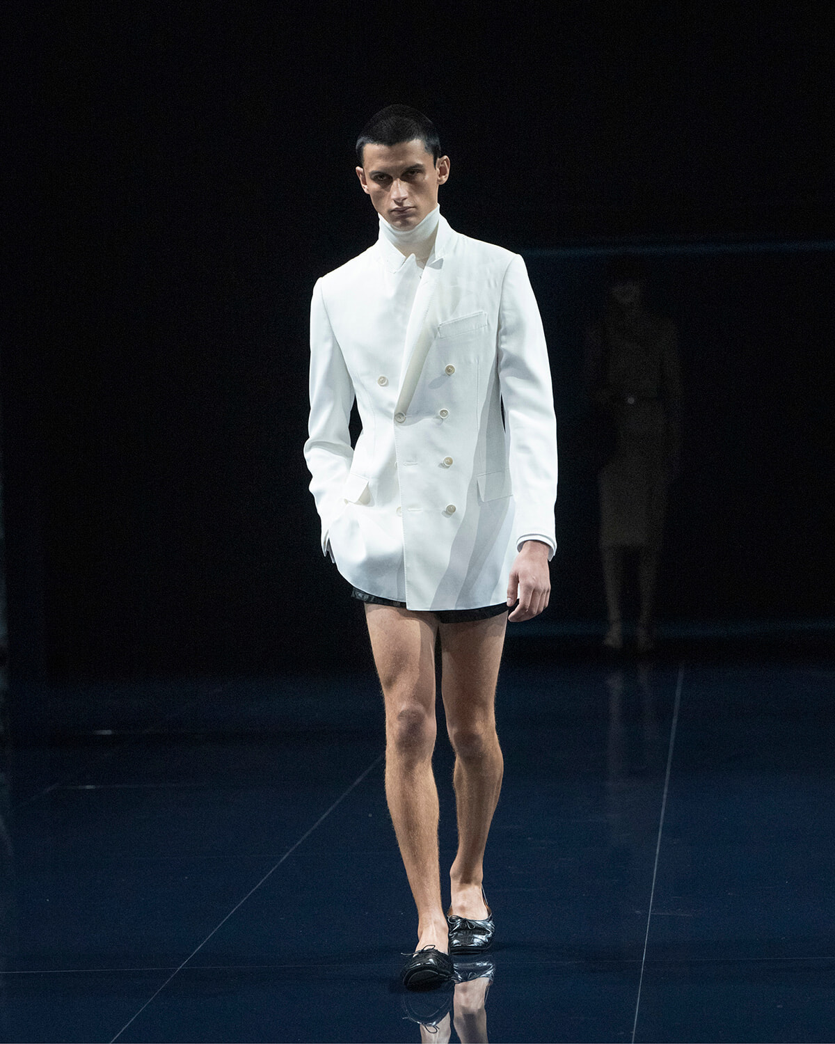 Male model walks a fashion runway wearing a white double-breasted blazer, white turtleneck, and black shorts on a dark stage.