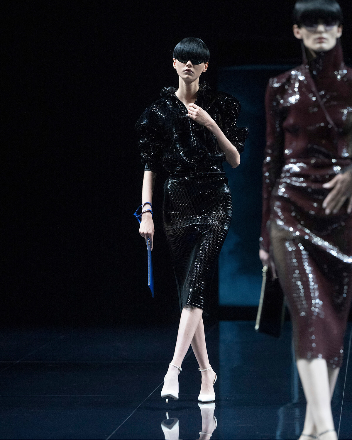 Runway model in a glossy black sequined outfit with voluminous sleeves, wearing sunglasses, and holding a blue clutch while walking the catwalk. Behind her, another model in a maroon sequined outfit is visible.