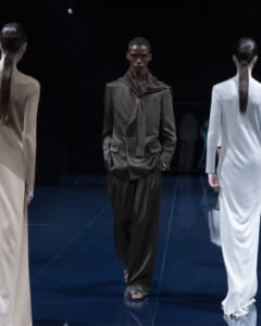 Male model in a dark, double-breasted suit with a long scarf walks a glossy runway, flanked by beige and white-clad models.