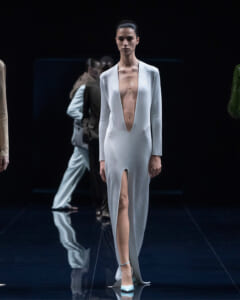 Model walks the runway in a pale blue, long-sleeve gown with a deep plunging neckline and high slit, on a dark stage.
