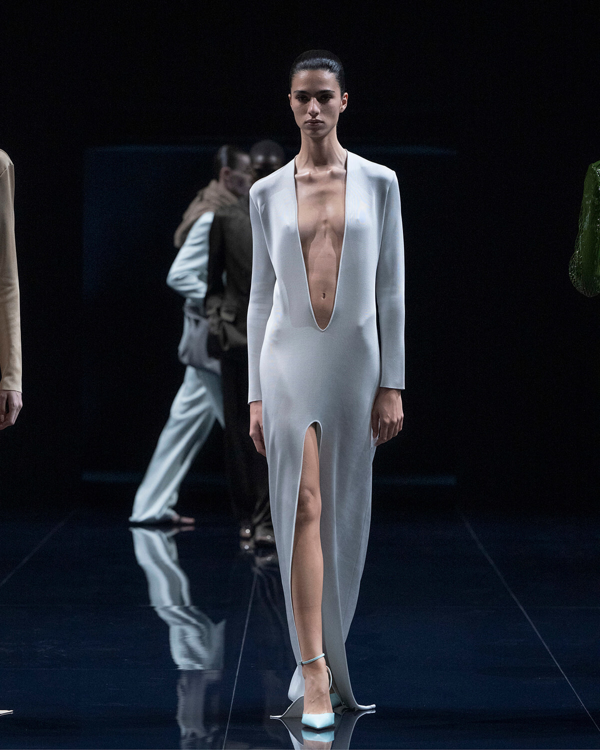 Model walks the runway in a pale blue, long-sleeve gown with a deep plunging neckline and high slit, on a dark stage.