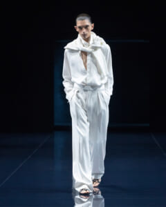 Male model in a white suit on a fashion runway, with a white scarf draped over his shoulders and hands in pockets.