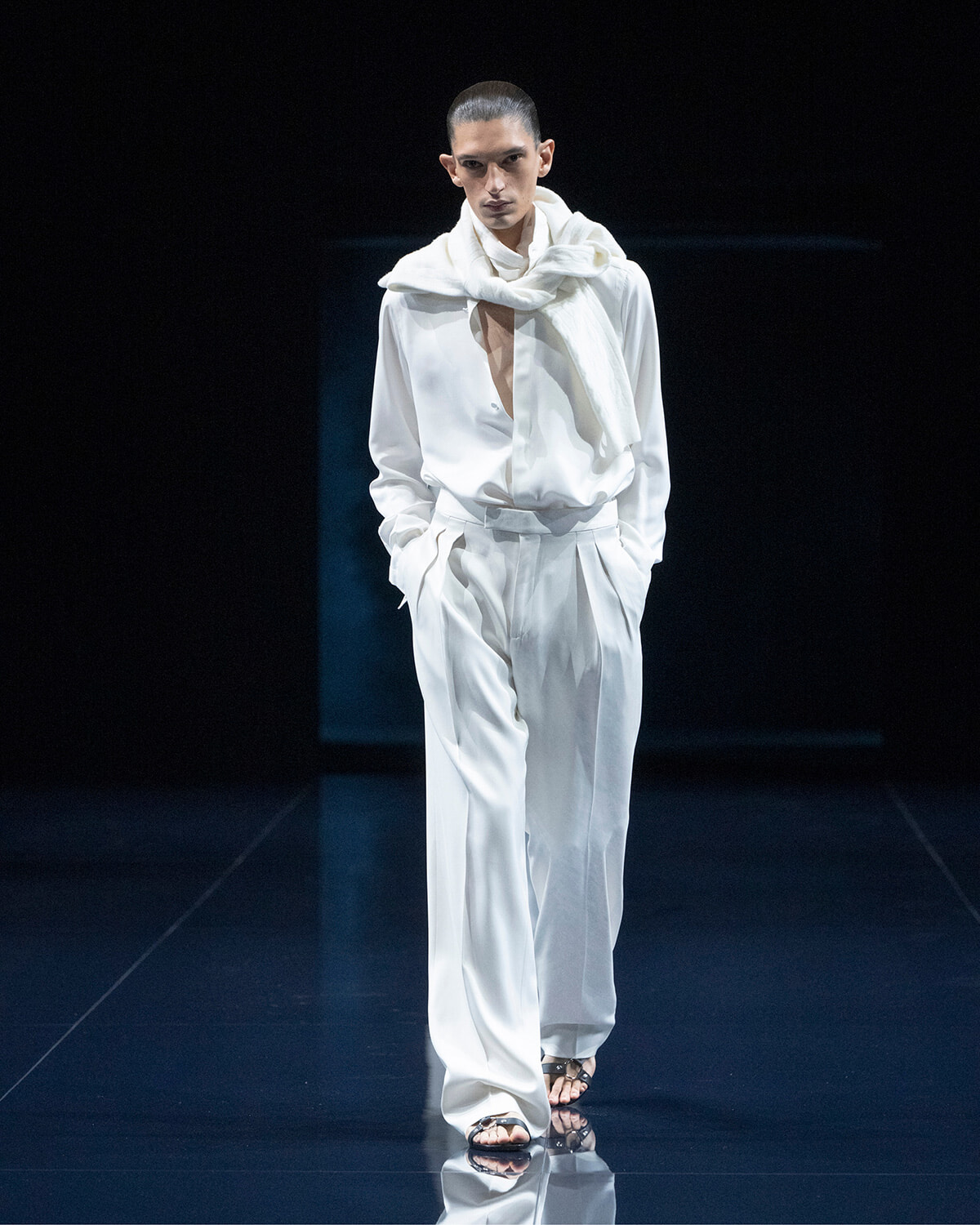 Male model in a white suit on a fashion runway, with a white scarf draped over his shoulders and hands in pockets.