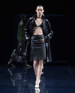 Model walks the runway in a black leather coat over a black bralette and lace-edged skirt, white heels.