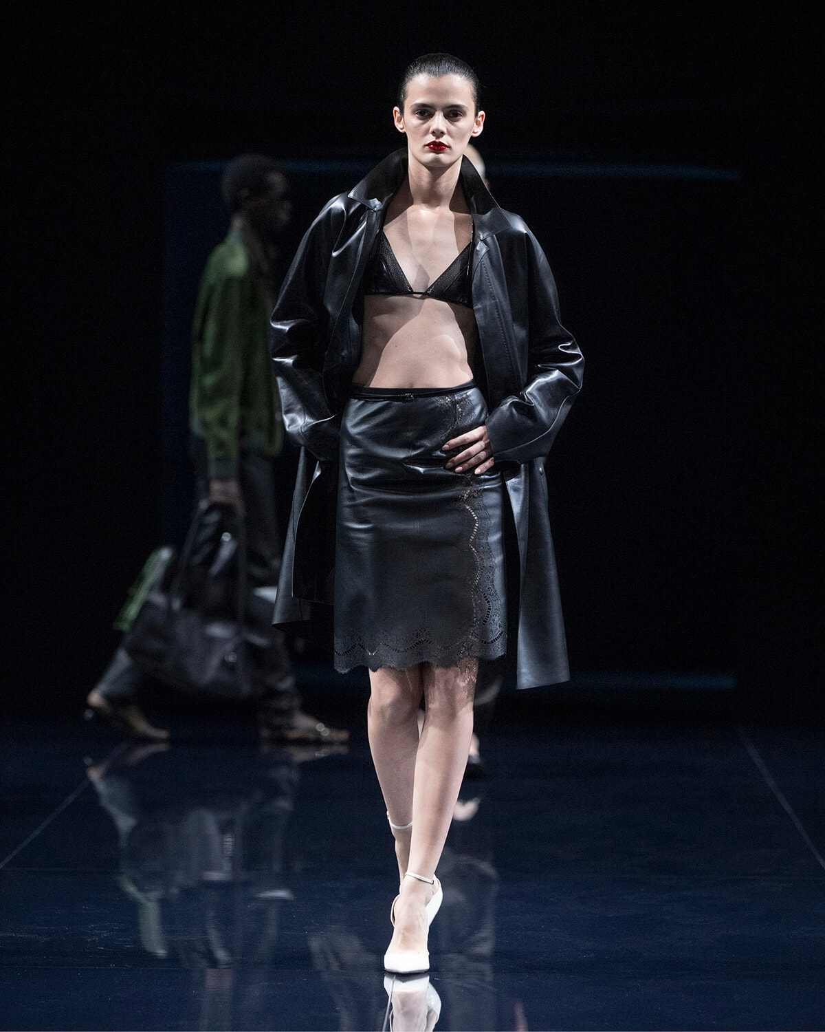 Model walks the runway in a black leather coat over a black bralette and lace-edged skirt, white heels.