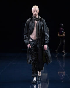 Male model walks a dark runway in a black raincoat, scarf, and layered black pants, hands in pockets.
