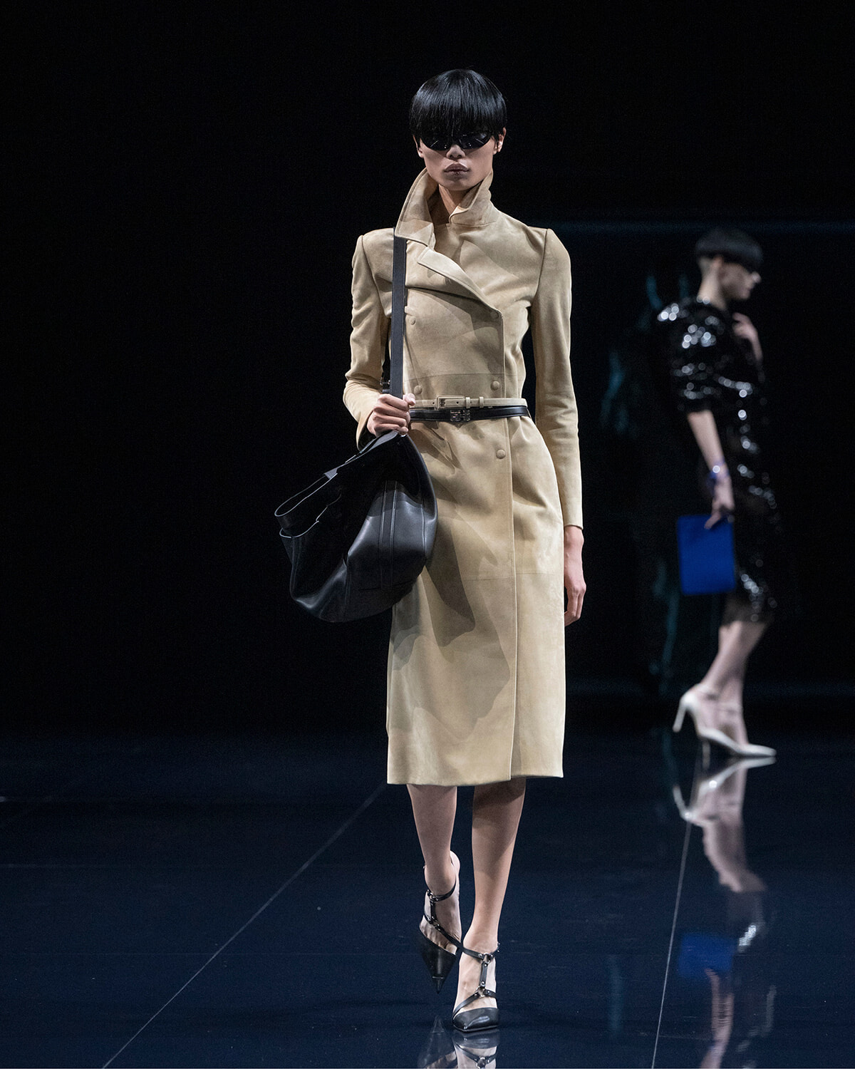 Runway model wearing a beige trench coat with a black belt, sunglasses, and a large black shoulder bag.