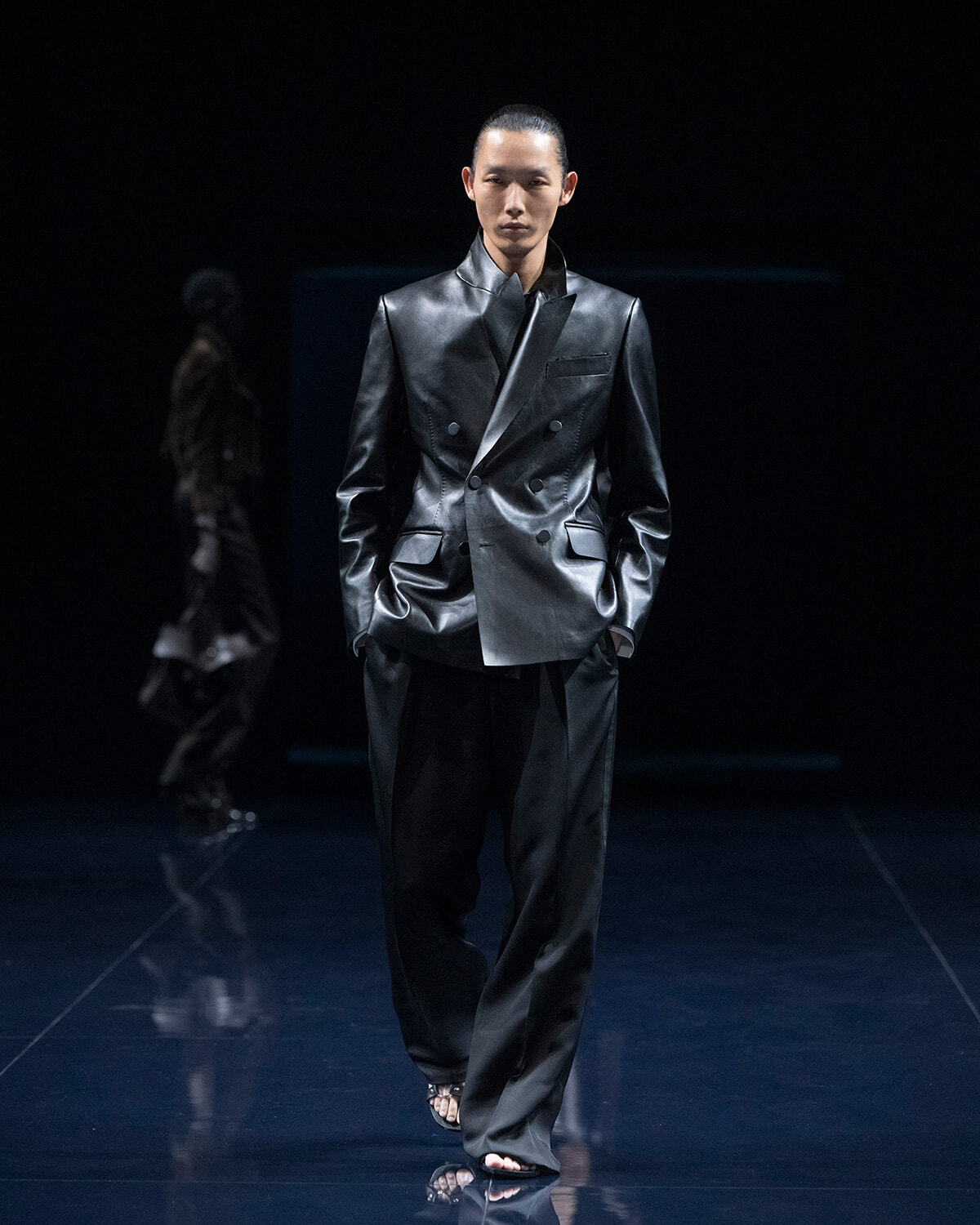 Male model walks a glossy black double-breasted suit on a dark runway.
