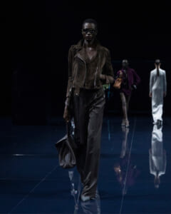 Model walks a dark runway wearing a brown suede biker jacket, dark trousers, sunglasses, and a brown bag.