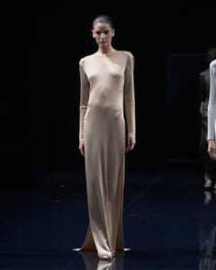 Model on a runway wearing a full-length beige satin dress with long sleeves and a neutral expression.