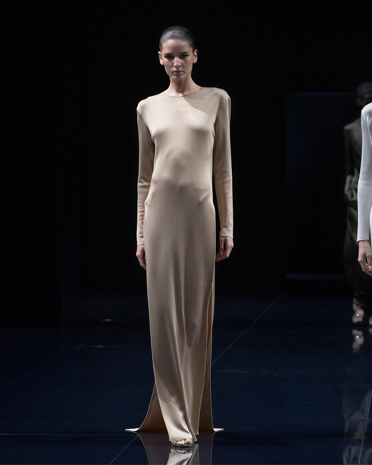 Model on a runway wearing a full-length beige satin dress with long sleeves and a neutral expression.