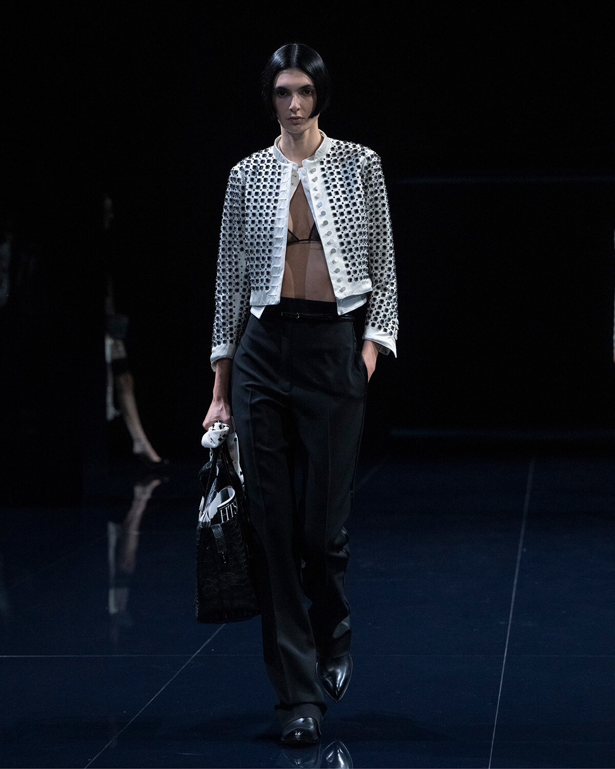 Model on a dark runway wearing a white studded cropped jacket over a sheer black top with black tailored pants, carrying a black handbag.