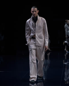 Male model walks a fashion runway wearing a light beige suit with an oversized jacket and patch pockets, against a dark backdrop.
