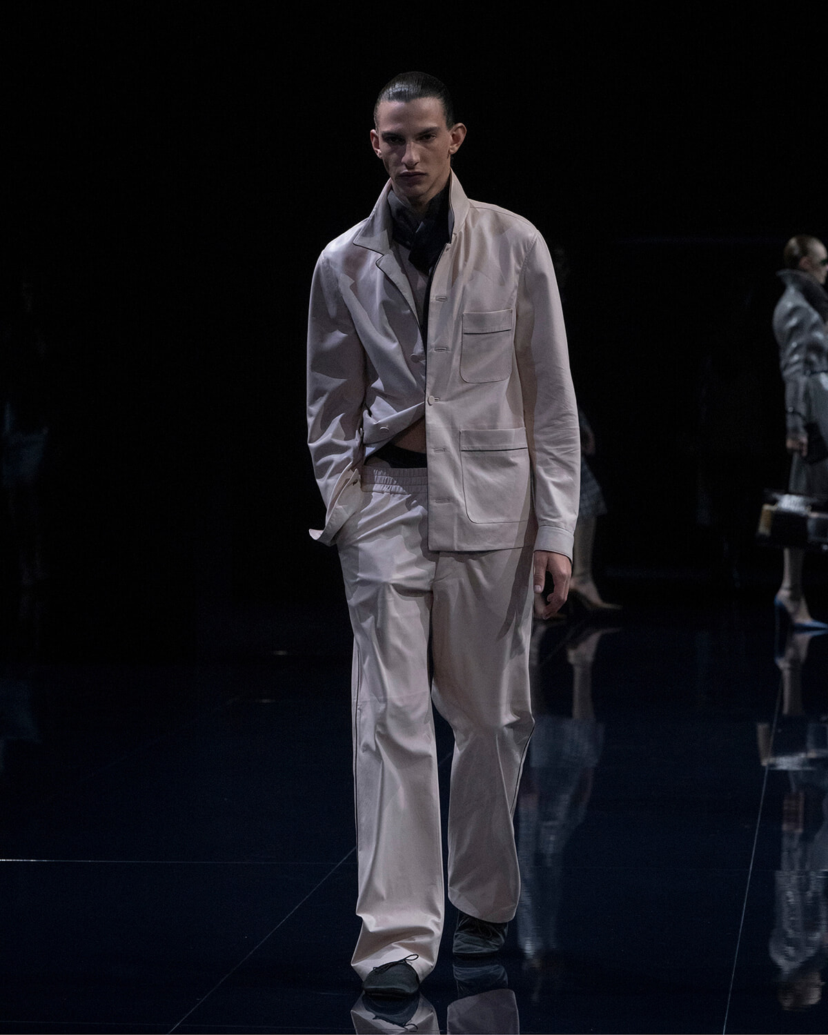 Male model walks a fashion runway wearing a light beige suit with an oversized jacket and patch pockets, against a dark backdrop.
