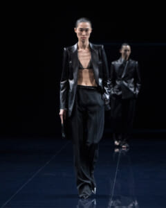 Model walks a dark runway in a black satin blazer, bralette, and wide trousers, holding a small clutch.