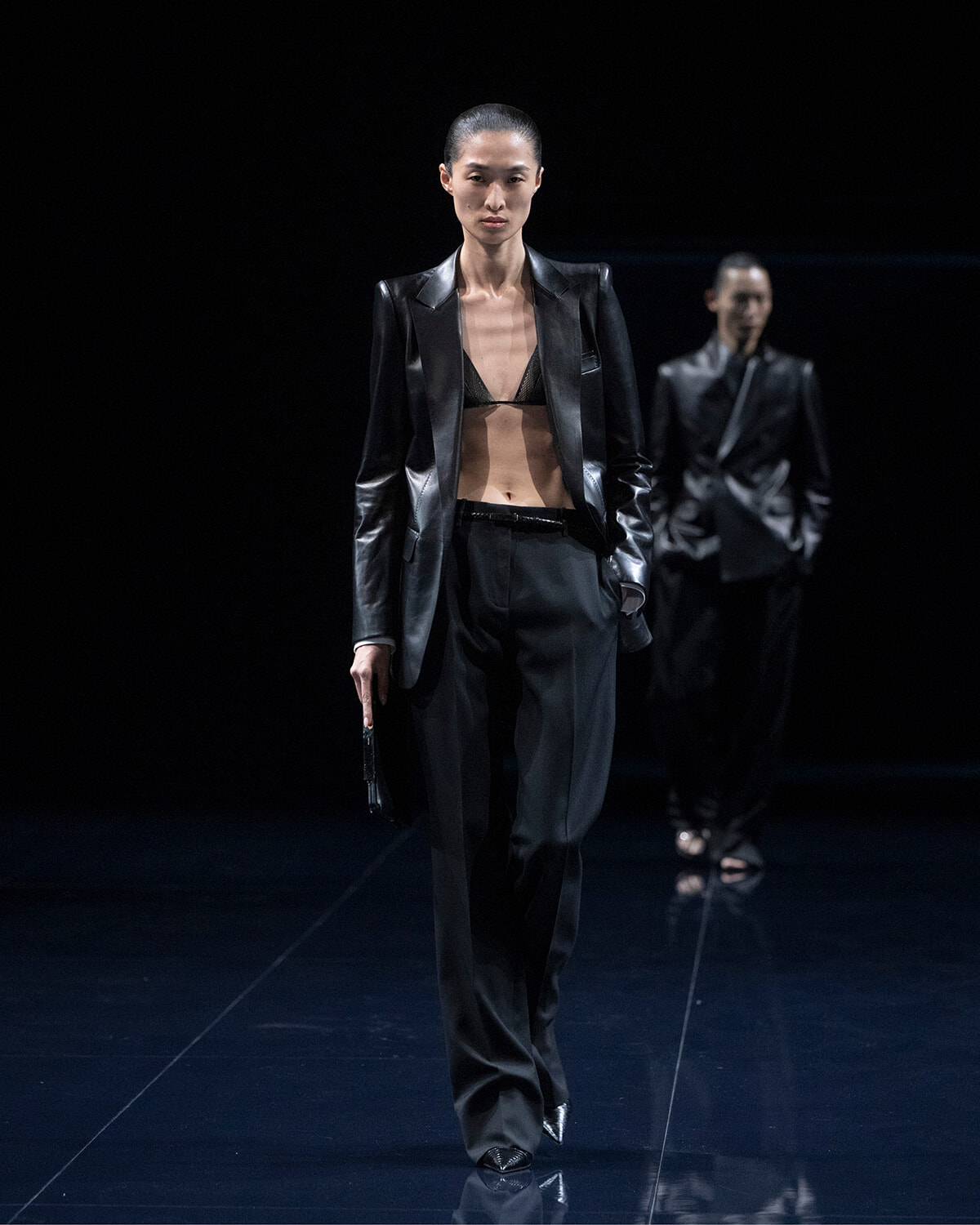 Model walks a dark runway in a black satin blazer, bralette, and wide trousers, holding a small clutch.