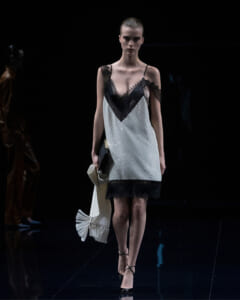 Model walking a fashion show in a black lace-trim slip dress with white polka-dot fabric, carrying a fringed scarf.