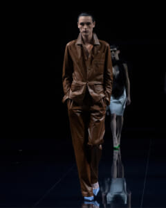 Model on a dark runway wearing a brown leather suit with a matching jacket and pants, hands in pockets.