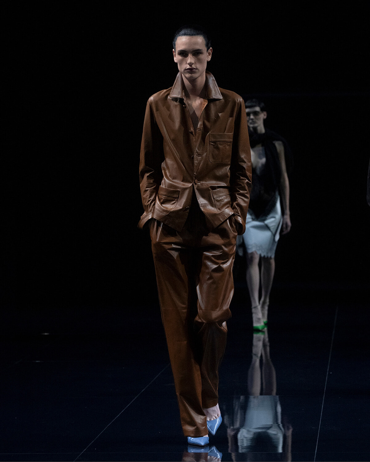 Model on a dark runway wearing a brown leather suit with a matching jacket and pants, hands in pockets.
