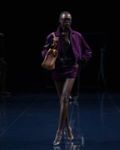 Runway model wearing a purple velvet blazer and matching shorts with a black top and tan handbag, sunglasses, walking on a dark catwalk.