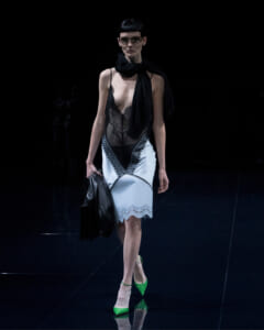 Runway model wearing a sheer black lace top over a pale blue lace-trimmed dress with lime-green heels and a black scarf, holding a black bag.