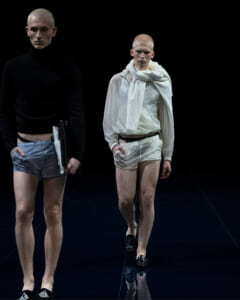 Two male models walk a dark runway; left in black turtleneck and light blue shorts, right in white ensemble with scarf.