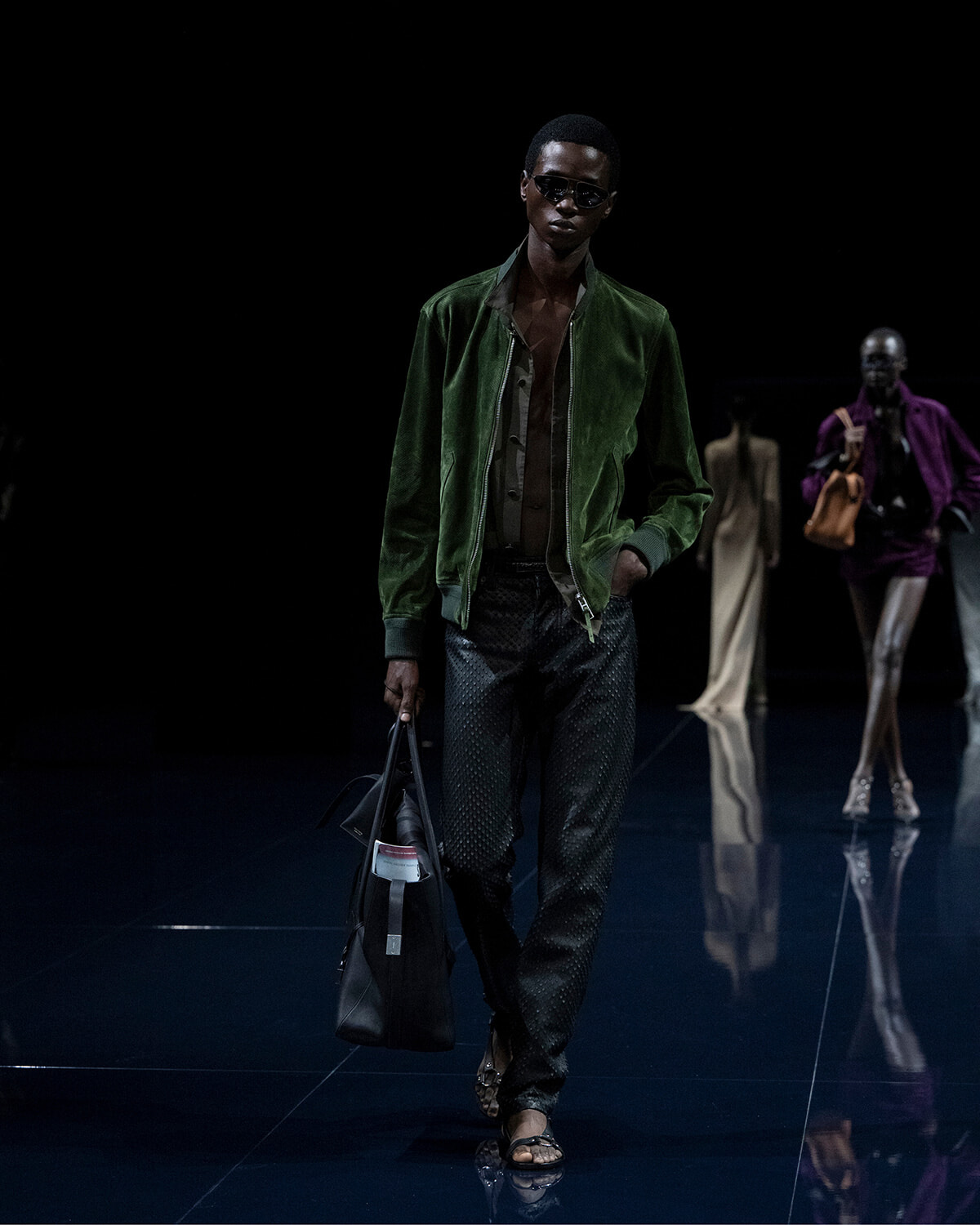 Male model walks a dark runway in a green bomber jacket, black patterned pants, sunglasses, and a black tote bag.