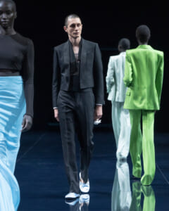 Male model in a black cropped blazer and matching pants walks the glossy runway; he holds a white cloth in his left hand.