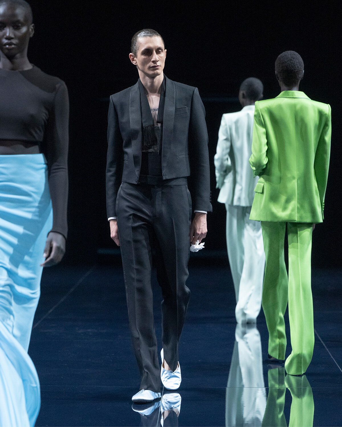 Male model in a black cropped blazer and matching pants walks the glossy runway; he holds a white cloth in his left hand.