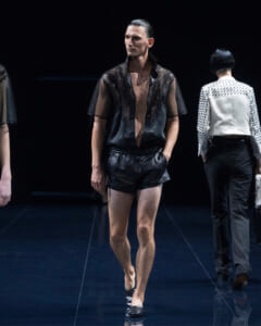 Male model walks the runway in a sheer black lace blouse and black satin shorts, sleek hair pulled back, dark stage setting.