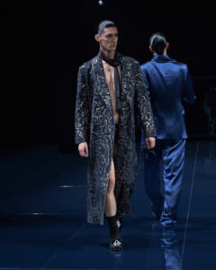 Male model on a dark runway wearing a long black patterned coat with a beige lining and a black scarf; another model in a blue satin suit facing away in the background.