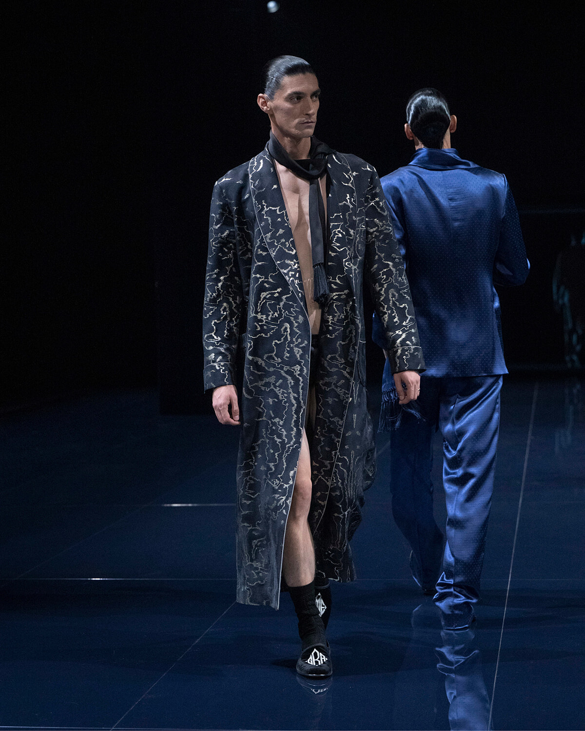 Male model on a dark runway wearing a long black patterned coat with a beige lining and a black scarf; another model in a blue satin suit facing away in the background.