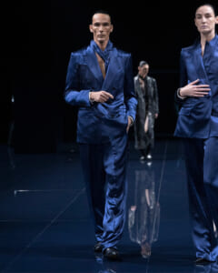 Male model in a blue satin double-breasted suit walking on a glossy runway, with another model in blue on the right and a dark background.
