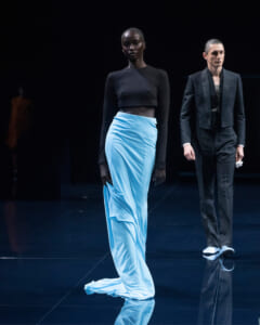 Model on a dimly lit runway wearing a black long-sleeve crop top and a flowing light-blue skirt; a man in a suit stands in the background, walking the catwalk.