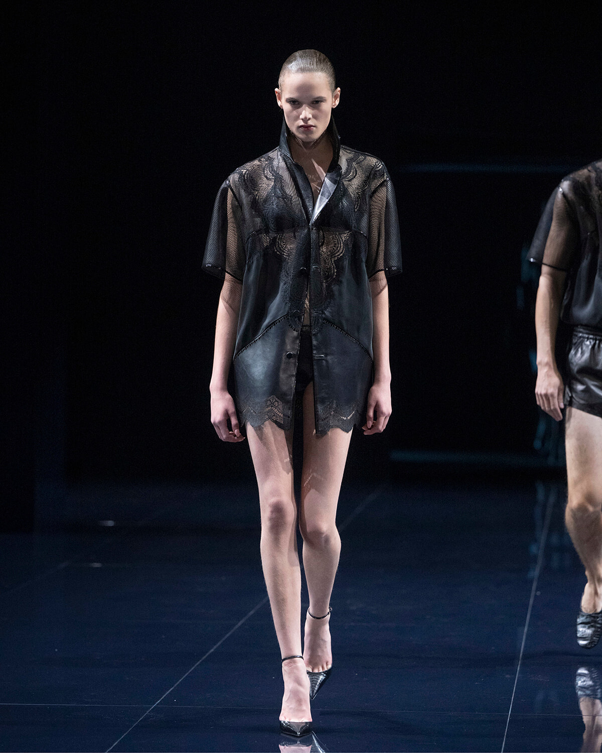 Model walks the runway in a sheer black lace shirt over a dark bra, paired with lace-trim shorts and pointed heels on a dark catwalk.